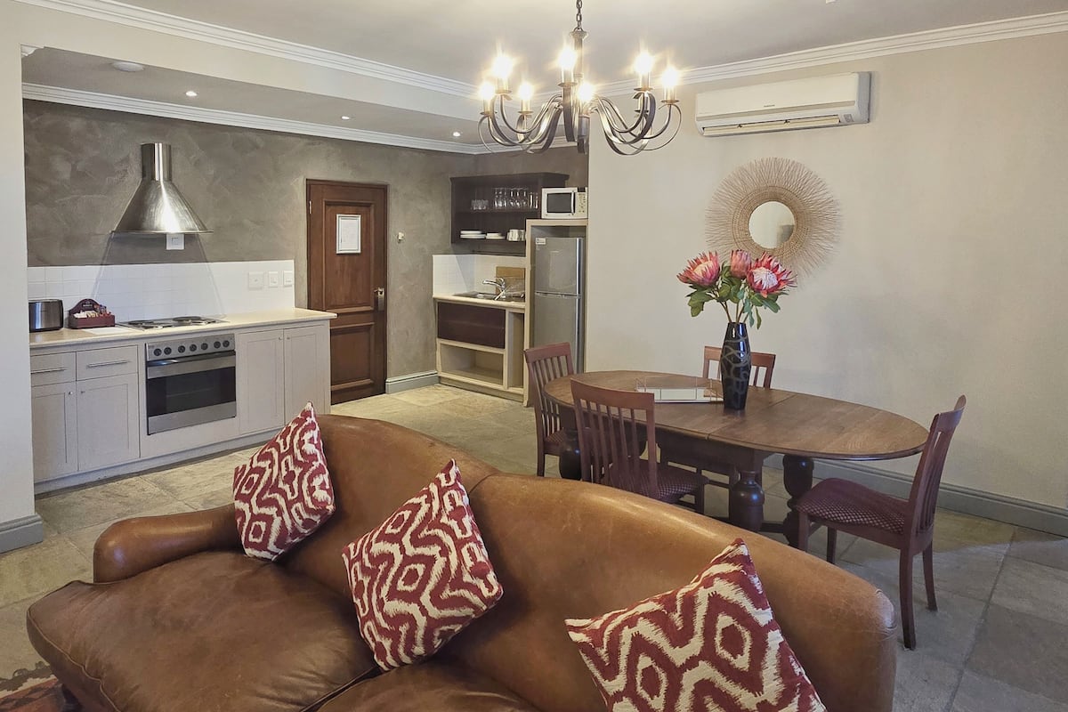 A modern kitchen with a gas stove, oven, and stainless steel range hood. There is a dining area with a wooden table and chairs, and a brown leather sofa with patterned cushions.