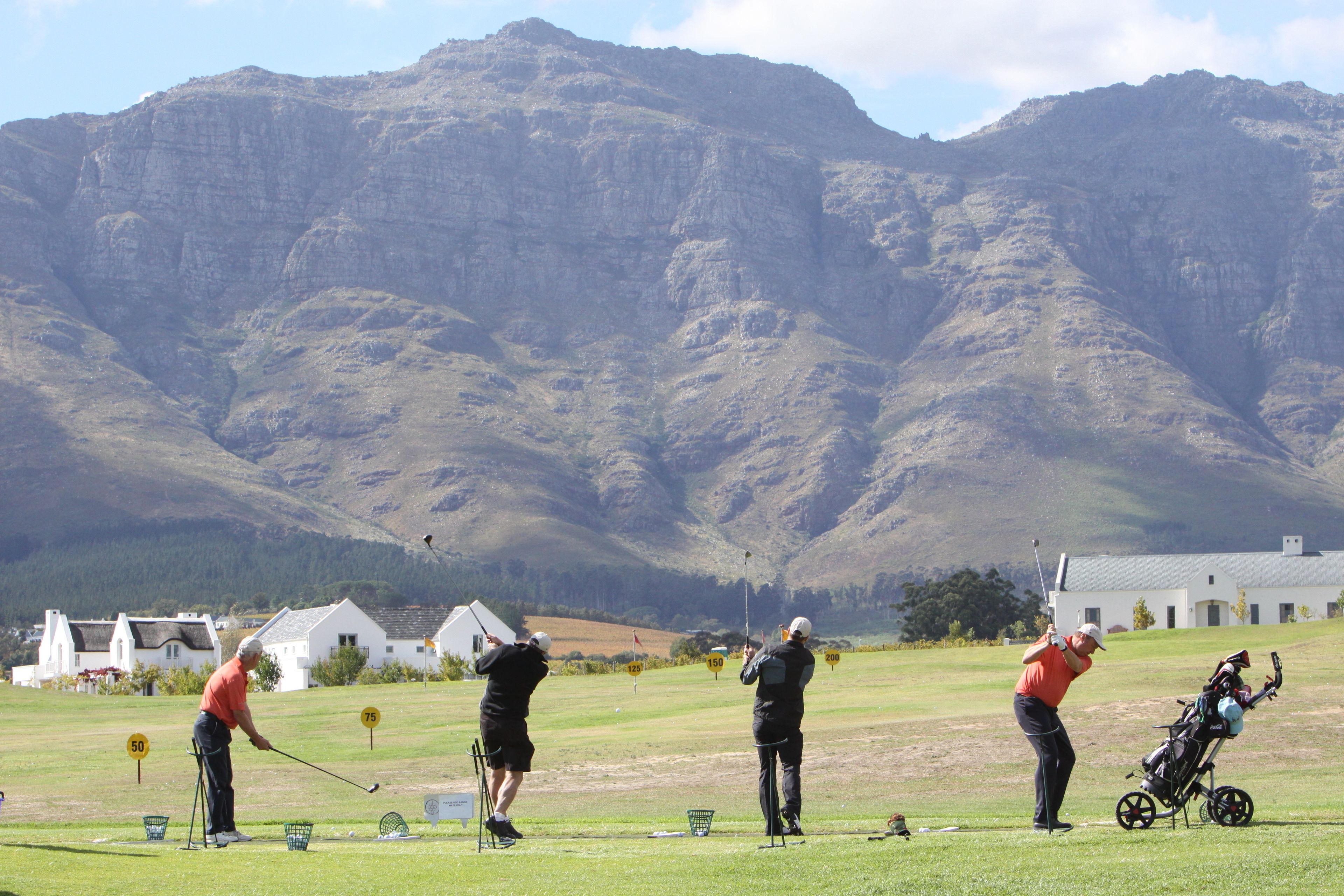 A golf course with players and a golf cart, surrounded by mountains and houses.