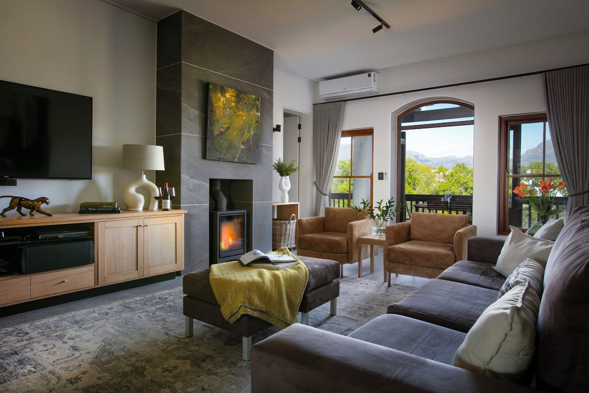 A modern living room with a fireplace, comfortable seating, a flat-screen TV, and a scenic view through large windows.