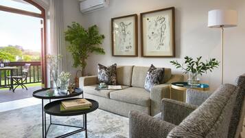 A living room with a sofa, armchairs, a coffee table, and a side table with a vase. There are two framed artworks on the wall, a potted plant, and a balcony with a view.