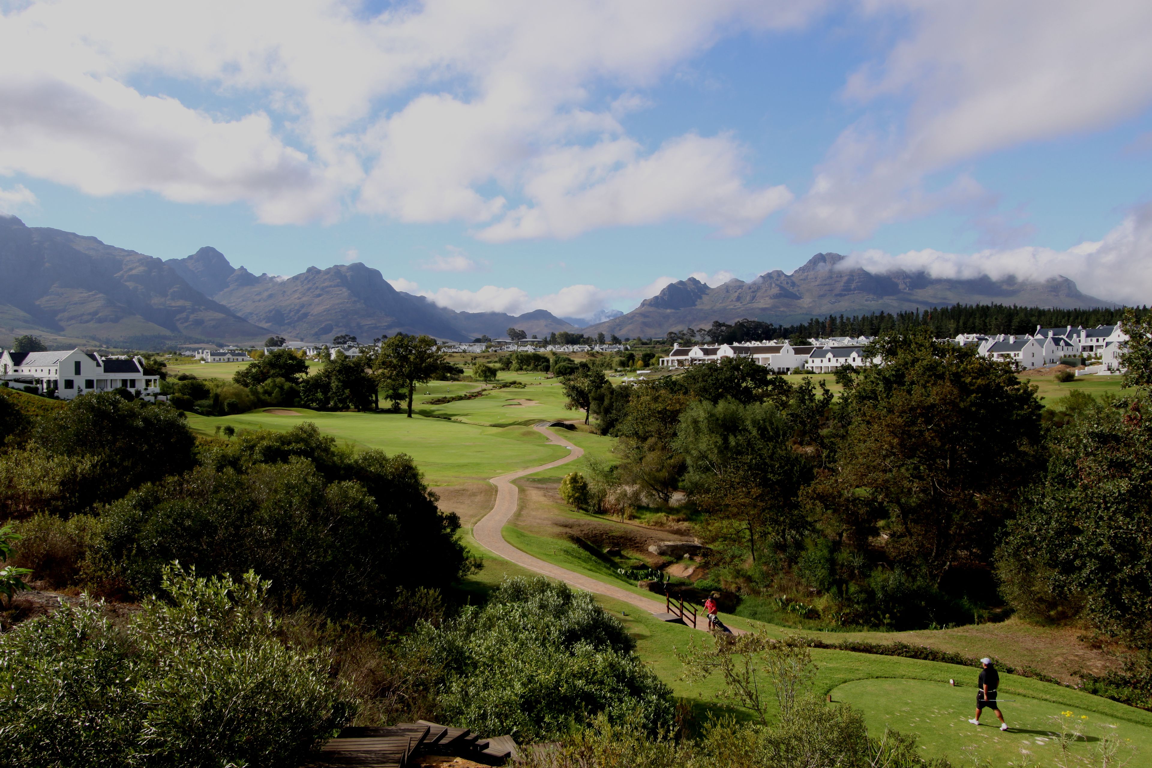 A golf course with a winding path, surrounded by greenery and mountains in the background.