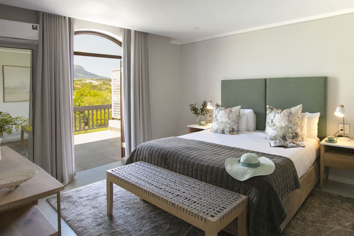 A hotel room with a large bed, a bench, a desk, and a balcony with a view of greenery and a mountain.