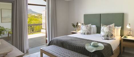 A hotel room with a large bed, a bench, a desk, and a balcony with a view of greenery and a mountain.