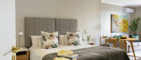 A neatly arranged bedroom with a bed, bedside tables, a small table with a bowl of fruit, and a painting on the wall.