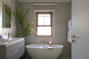 Ein modernes Badezimmer mit einer freistehenden Badewanne, einem Waschtisch mit Spiegel und einem Fenster mit Jalousien.