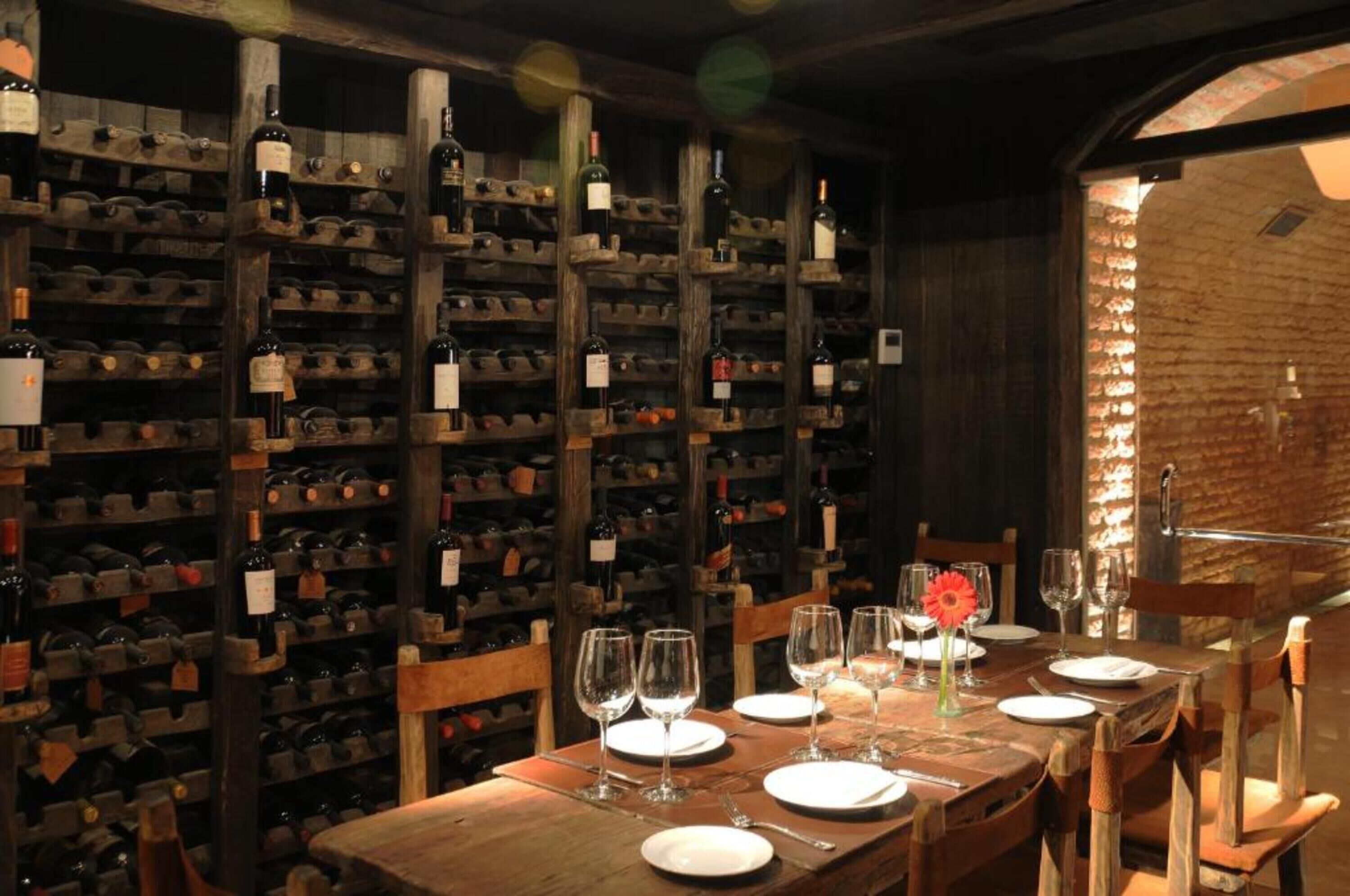 A wine cellar with wooden racks of wine bottles, a dining table set with wine glasses and plates, and a brick wall.