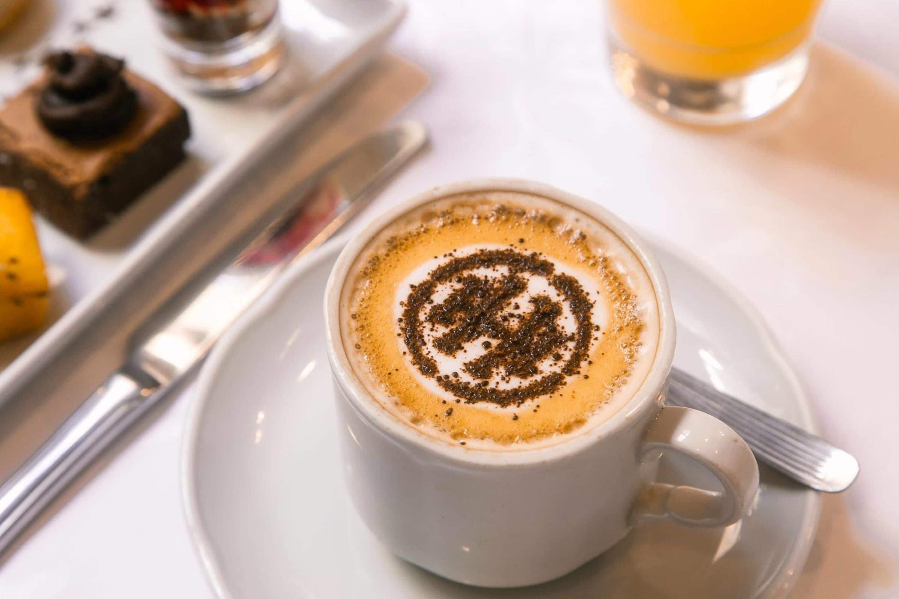 Una taza de café con arte de latte sobre un plato, una cuchara y un vaso de zumo de naranja en una bandeja.