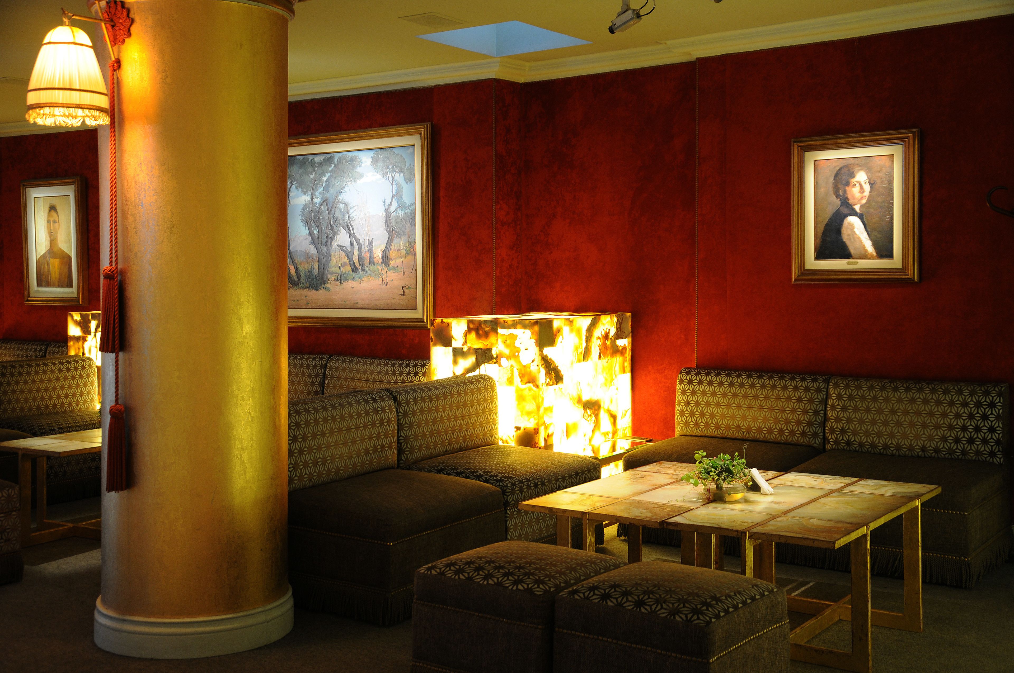 A room with red walls, a large gold column, and a fireplace with a painting above it. There are seating areas with patterned cushions and a table with a plant.