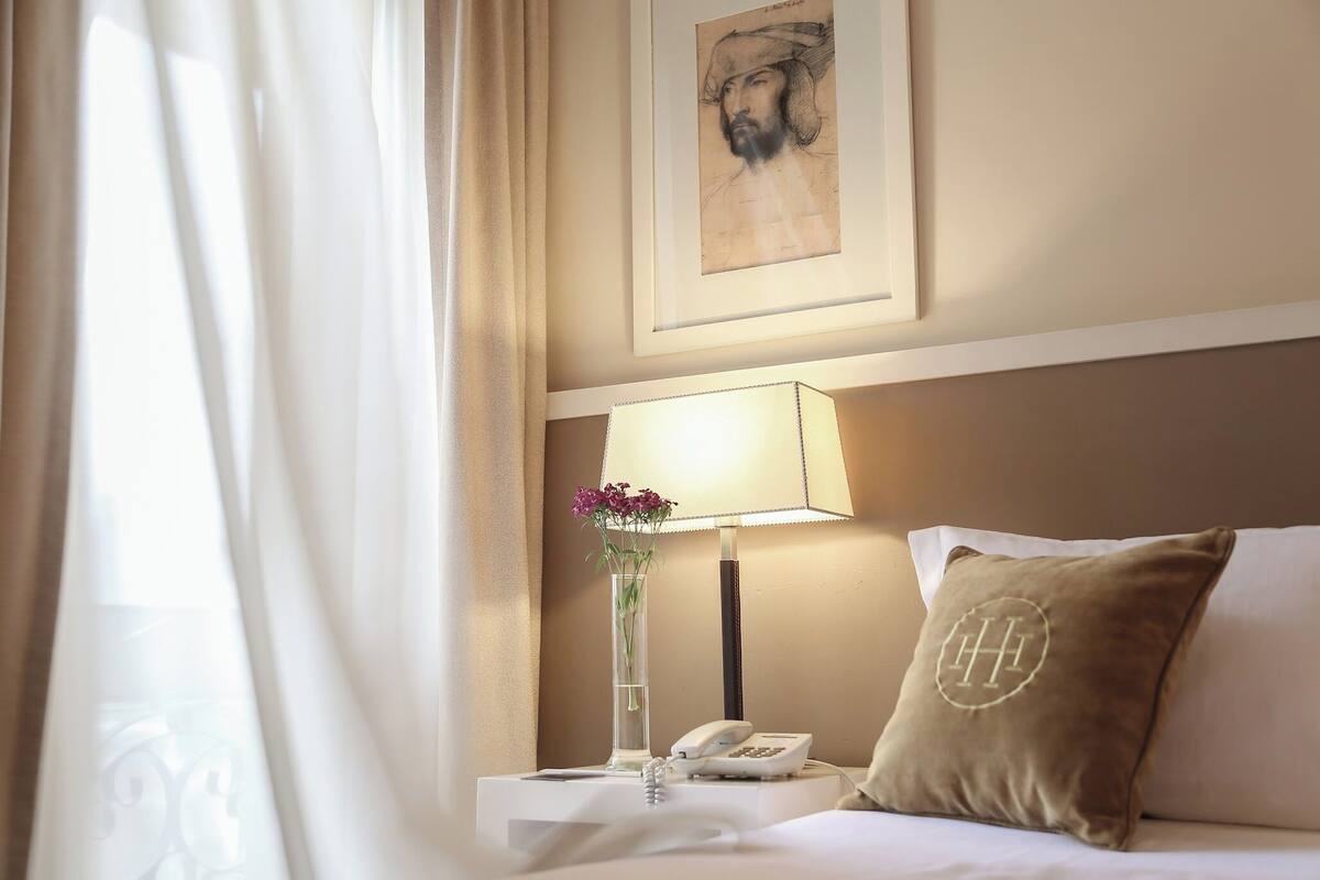 A hotel room with a bed, a bedside table with a lamp, a vase with flowers, a framed artwork on the wall, and a pillow with a monogram.