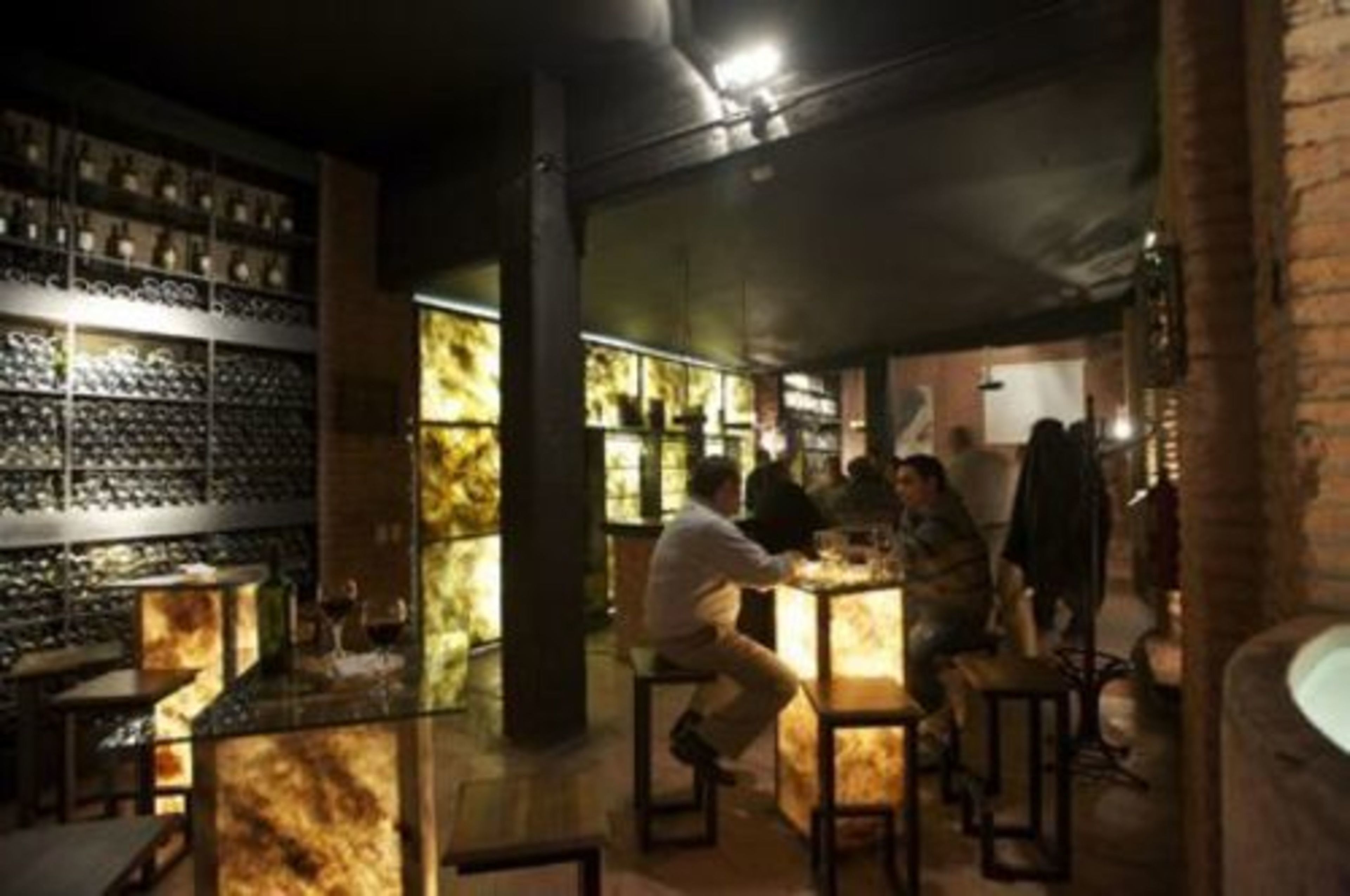 A bar area with patrons seated at tables, shelves with bottles, and a brick wall.