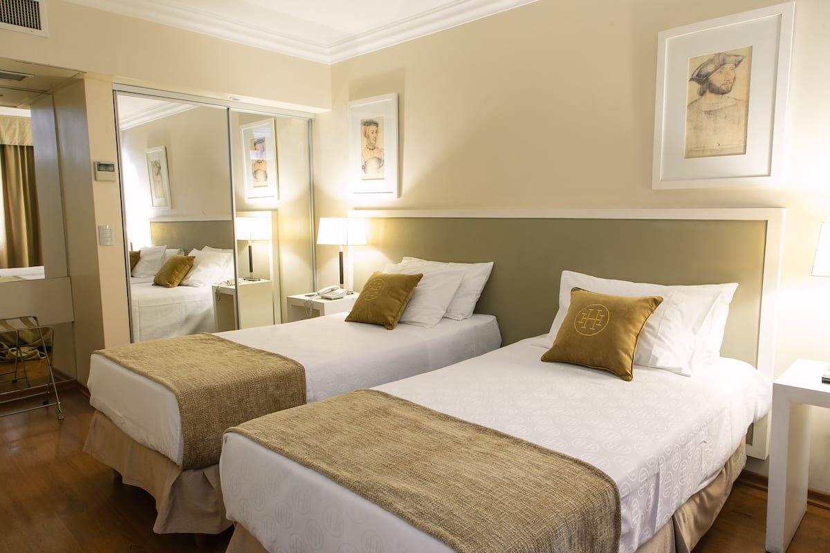 A hotel room with two beds, a large mirror, bedside tables with lamps, and artwork on the walls.