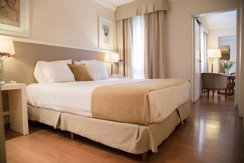 A hotel room with a large bed, bedside tables, a chair, and a door leading to another room.