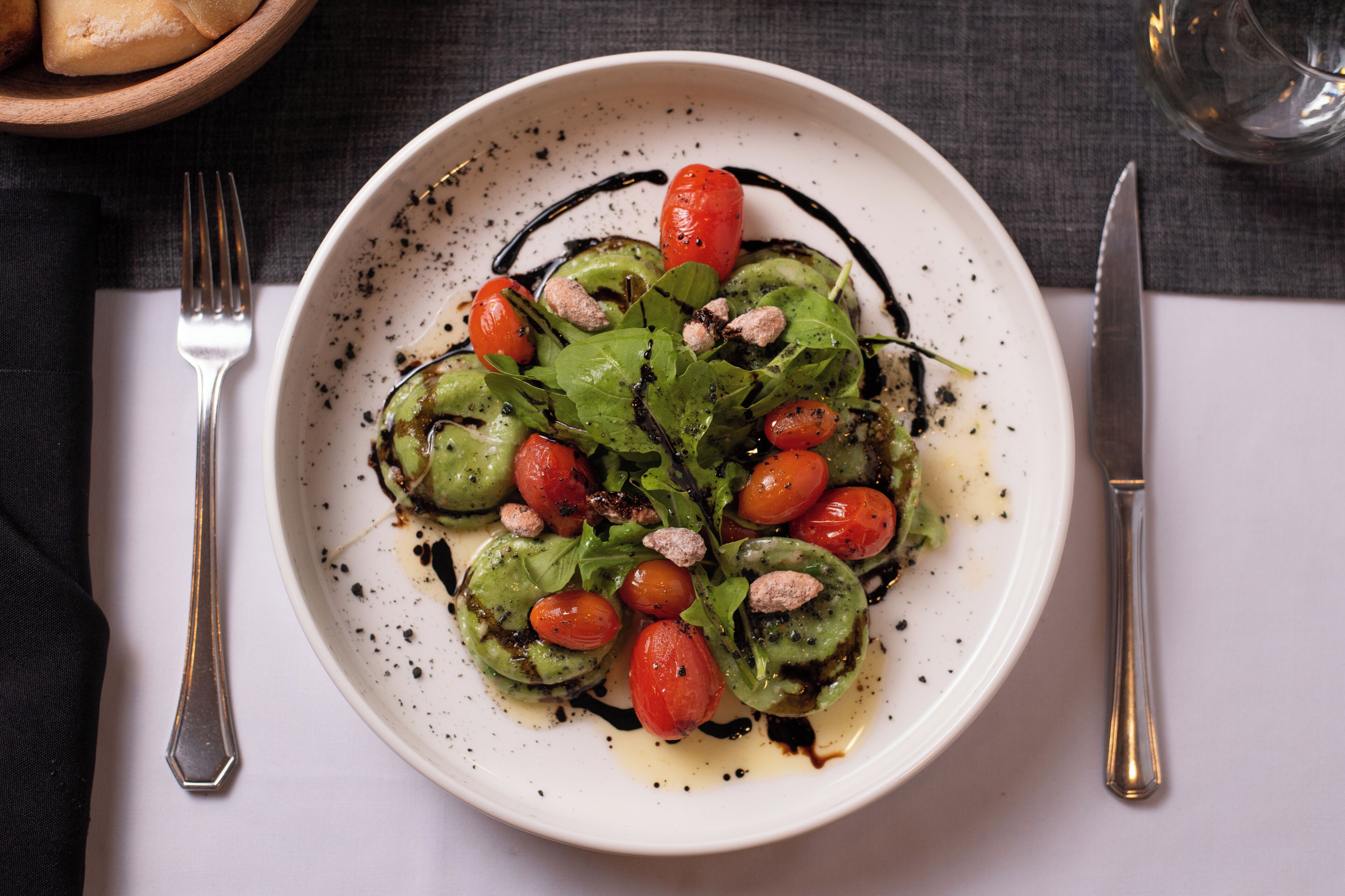 A white plate with a salad of greens, cherry tomatoes, and nuts, accompanied by a fork and knife on a dark cloth.
