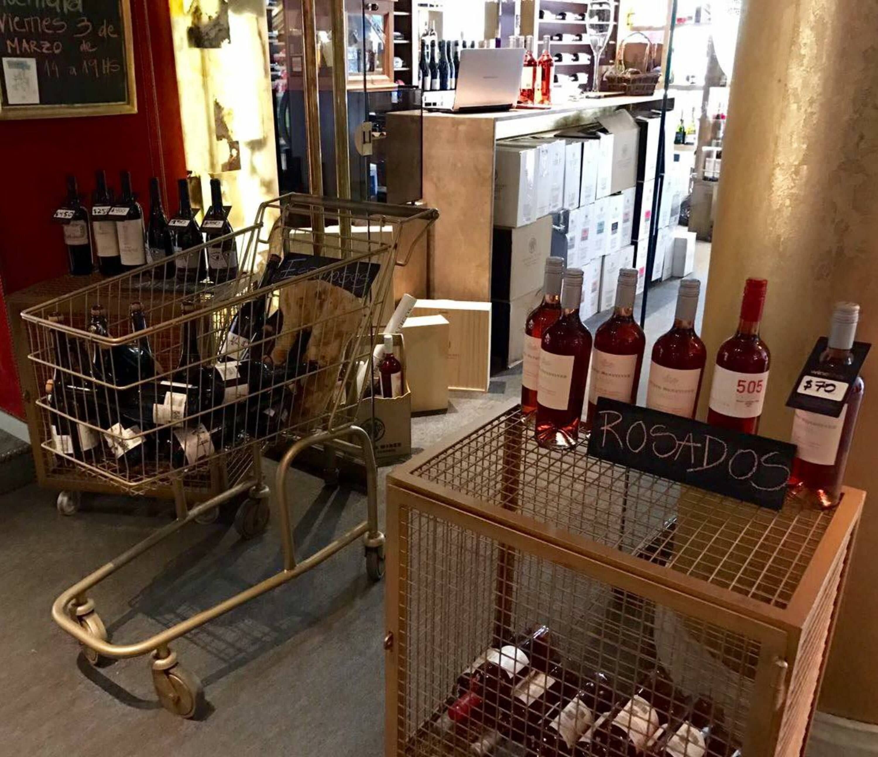 A wine shop interior with carts filled with wine bottles, a sign reading "ROGADOS", and a chalkboard with handwritten text.