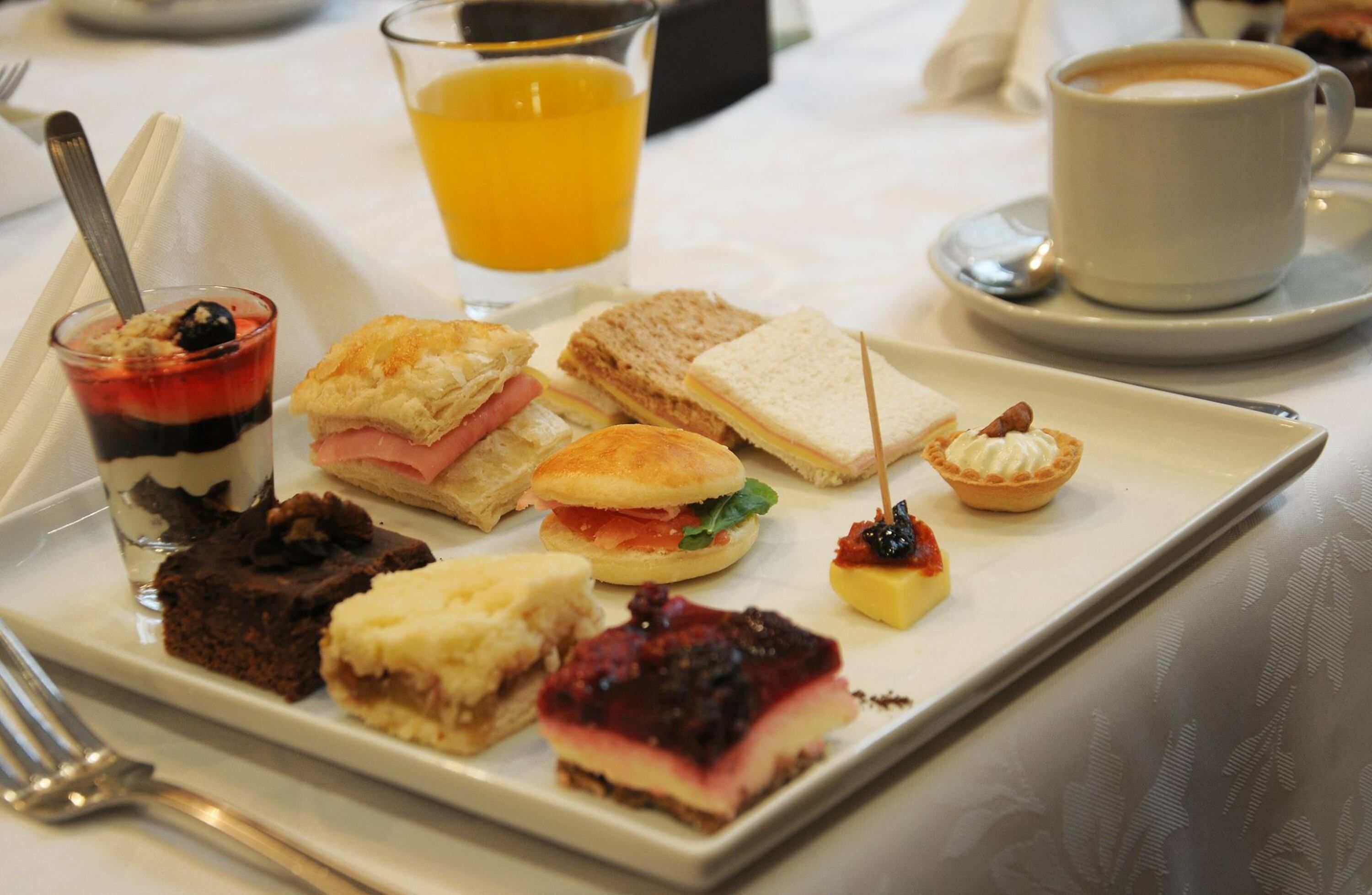 A variety of desserts and sandwiches on a white rectangular plate, accompanied by a glass of orange juice and a cup of coffee on a table.