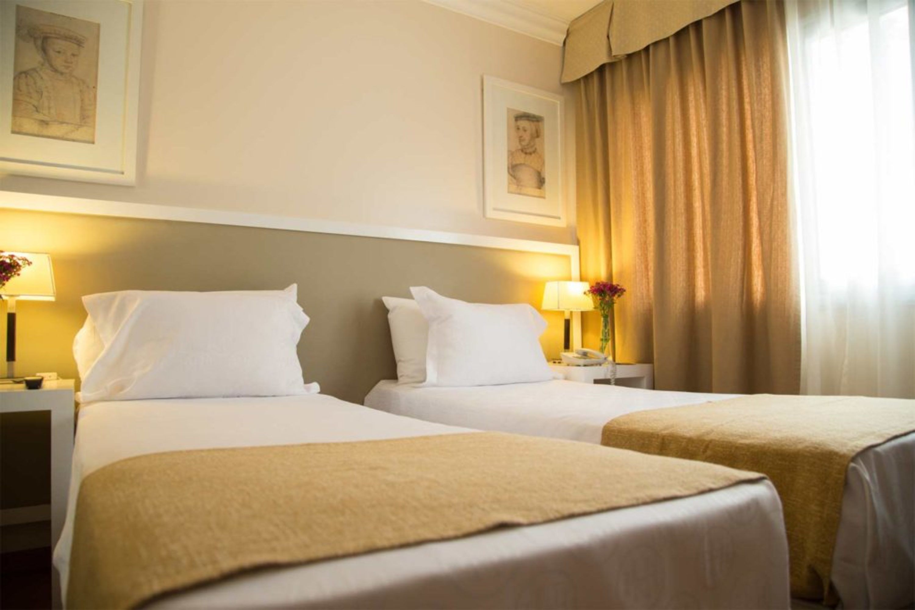 A hotel room with two beds, each with white linens and a beige throw blanket. There are bedside tables with lamps and a vase with flowers. The room has a neutral color scheme with beige walls and curtains.