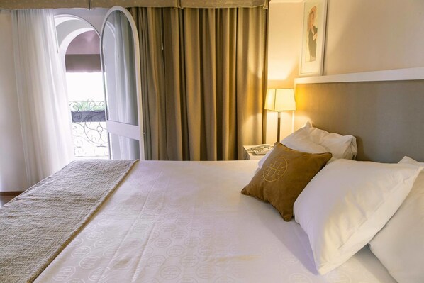 A hotel room with a large bed, a view of the outdoors through an arched window, and a lamp on the nightstand.