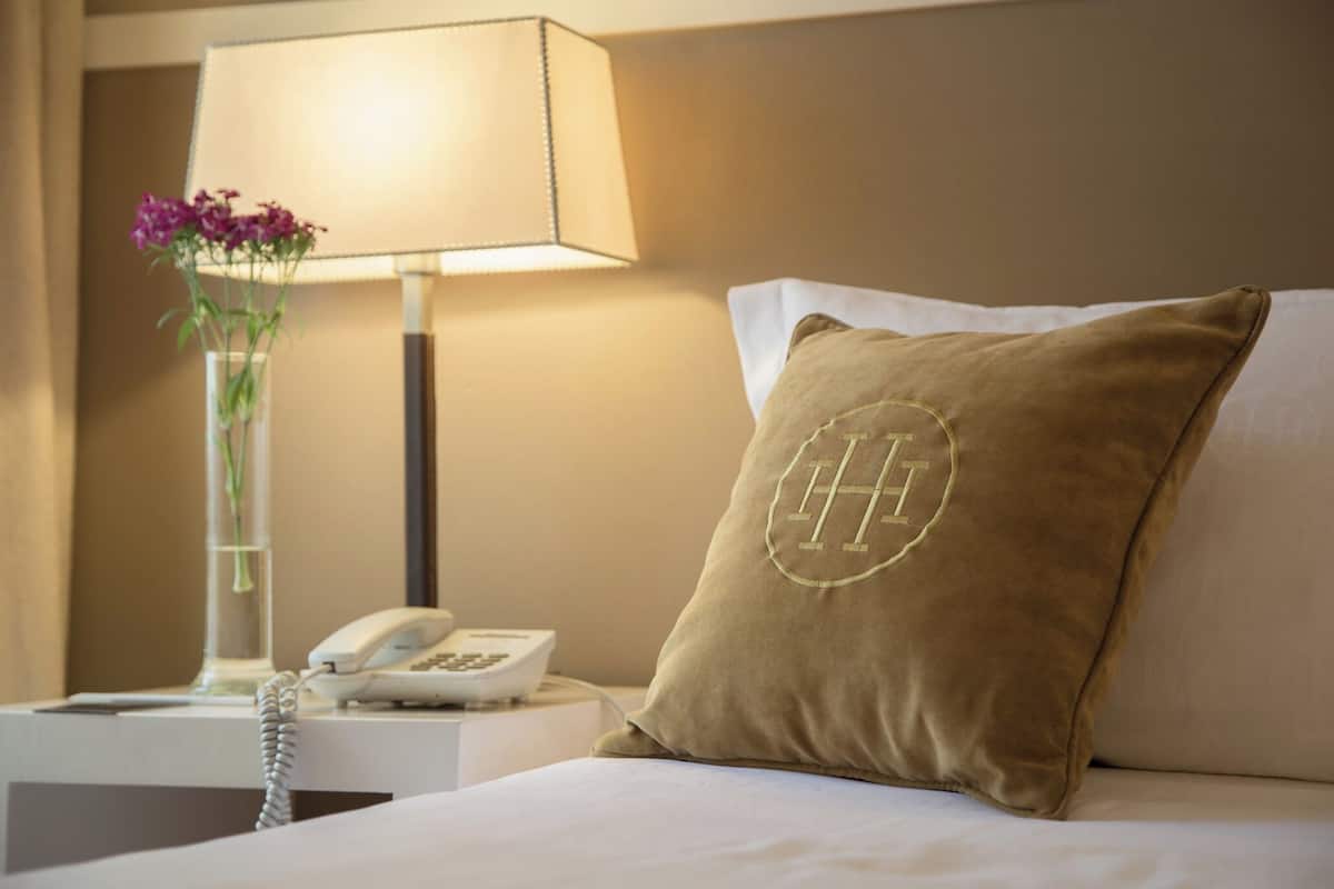 A hotel room with a bed, a nightstand with a lamp, a vase with flowers, and a phone.