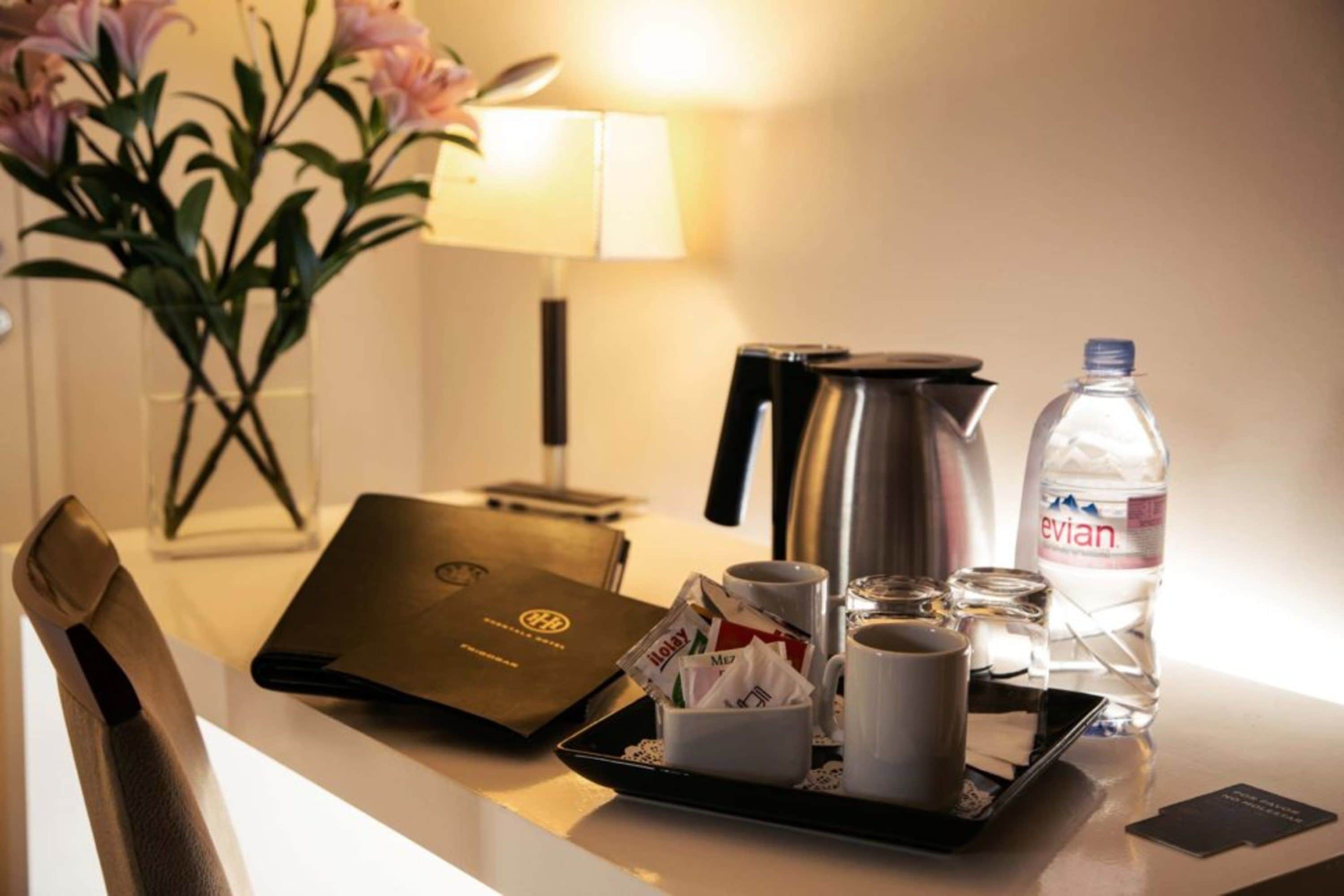 A hotel room with a desk featuring a laptop, a tray with coffee cups, a water bottle, and a vase with flowers.