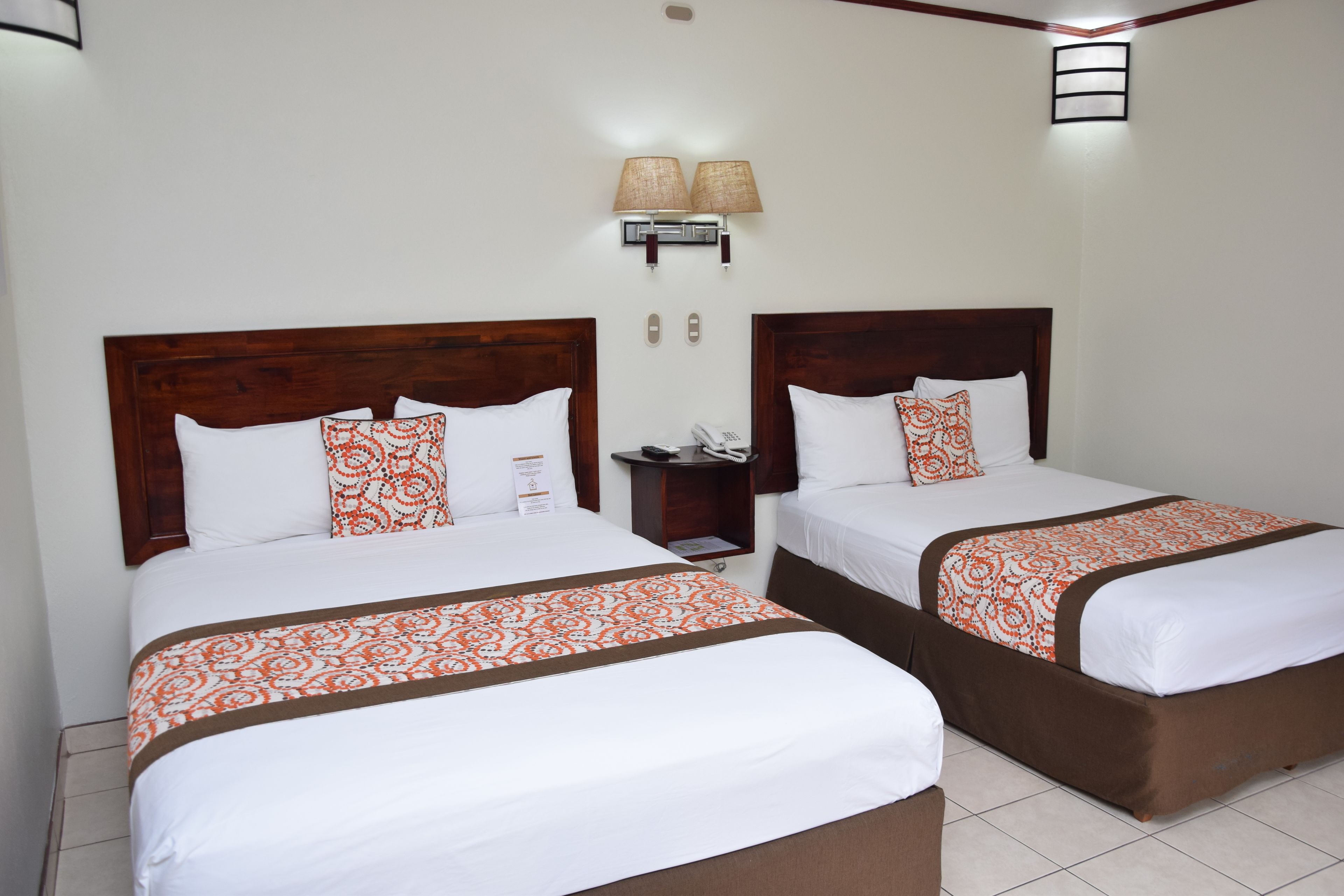 Two identical twin beds with white and orange patterned bedspreads, a wooden headboard, a nightstand with a lamp, and a wall-mounted light fixture.