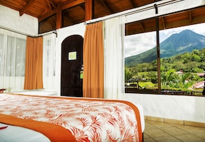 A bedroom with a bed, a door, and a window with a view of a mountain and greenery. - Arenal Volcano Inn (La Fortuna)