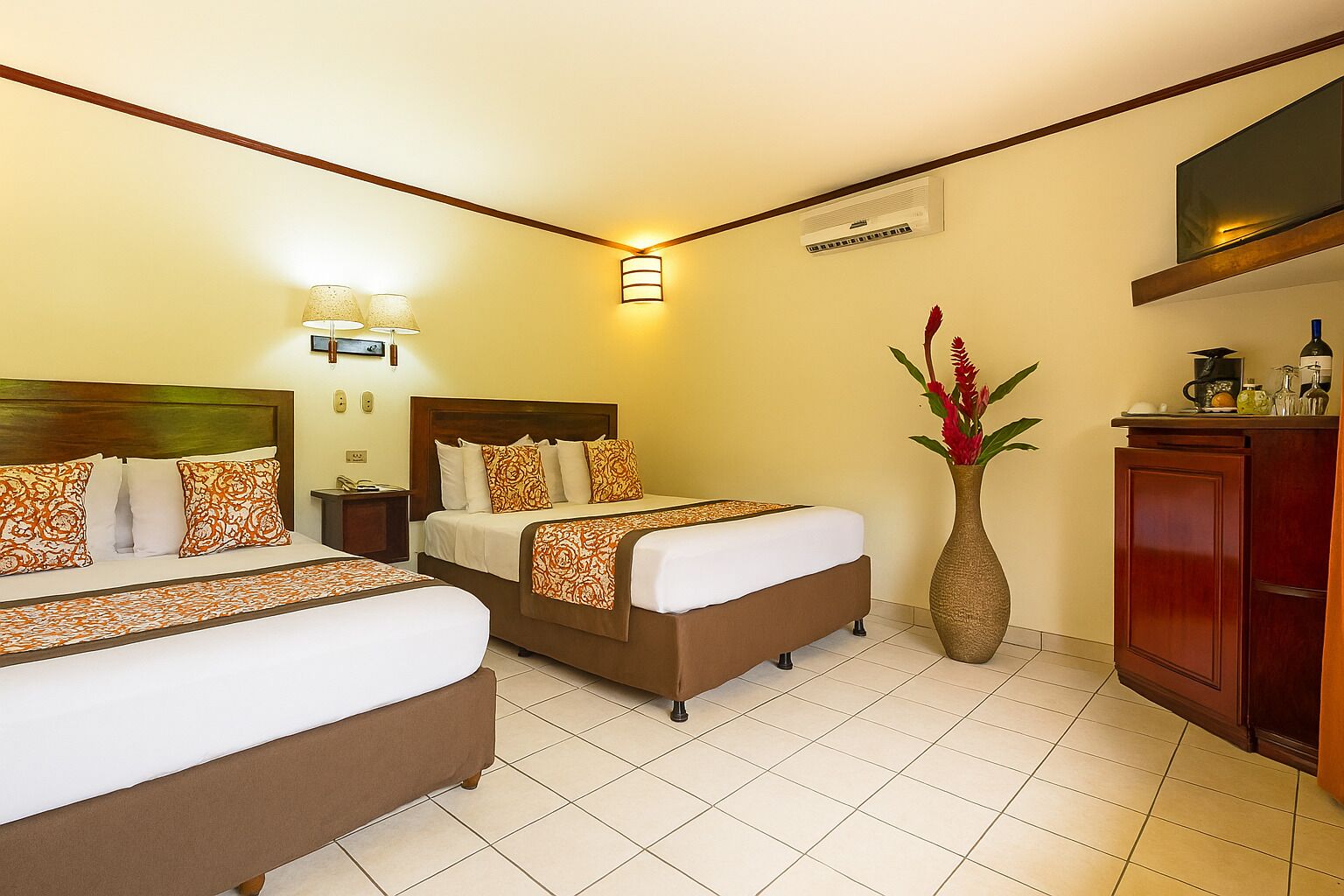 Une chambre d’hôtel avec deux lits, une table de chevet, une lampe, un vase avec des fleurs, une télévision et un minibar.
