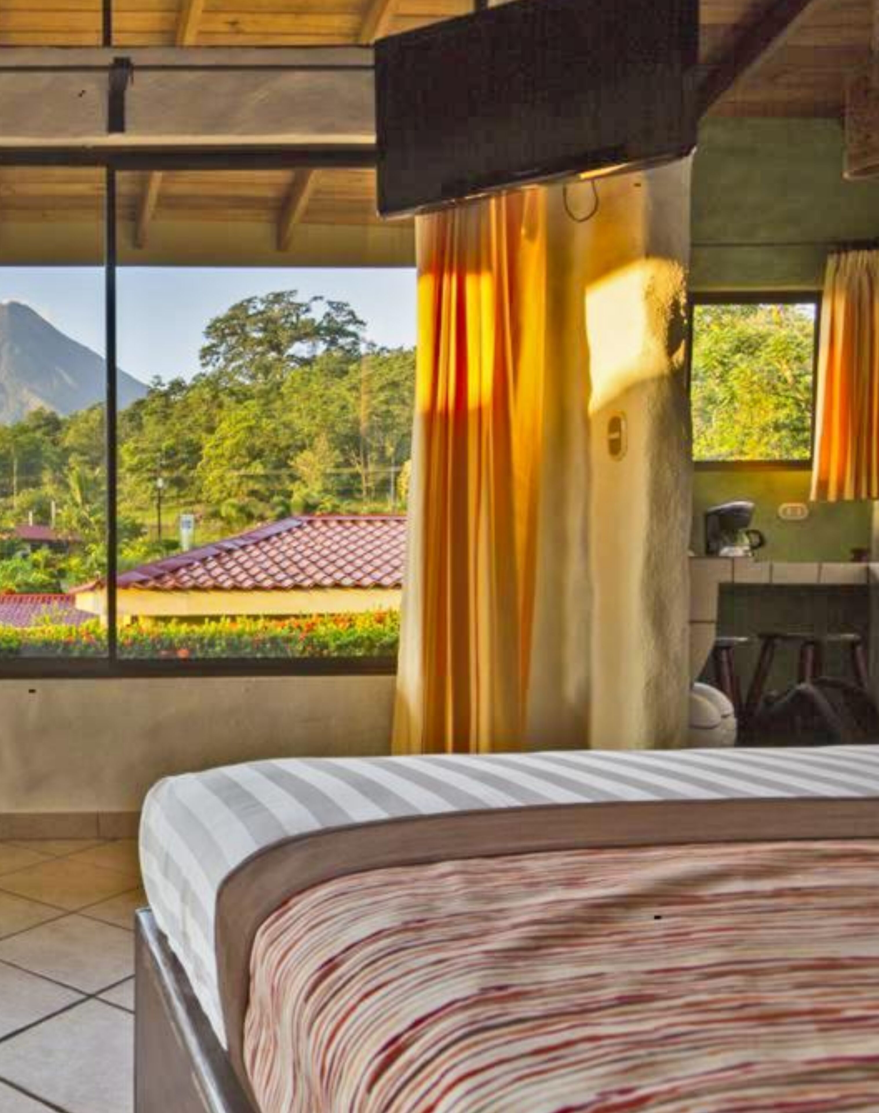 A hotel room with a bed, a window with curtains, and a view of a mountain and greenery.