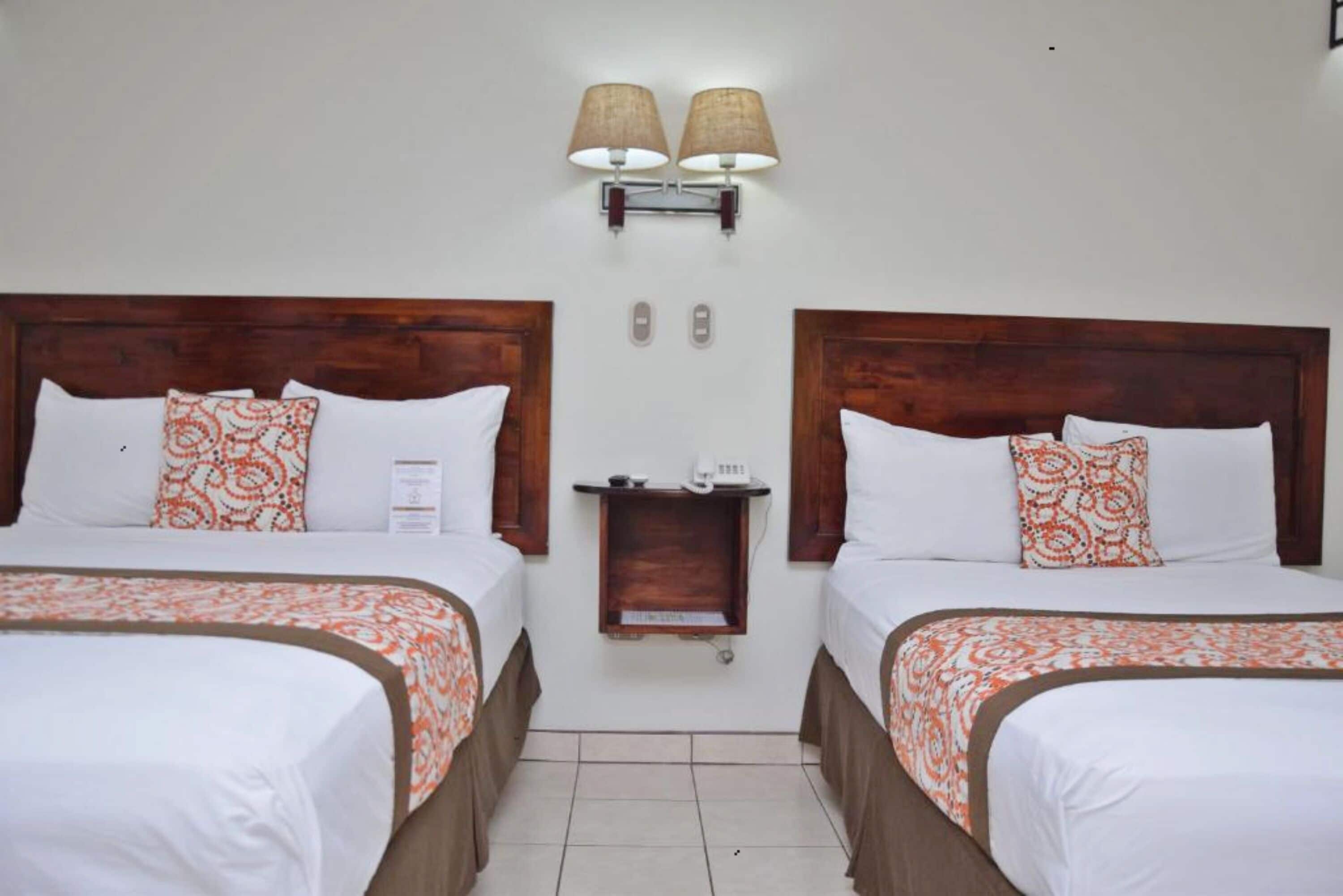 Habitación de hotel con dos camas, cada una con cabecero de madera, ropa de cama blanca y colchas estampadas en naranja. Hay dos lámparas de pared y un pequeño estante de madera con un teléfono y un cartel.