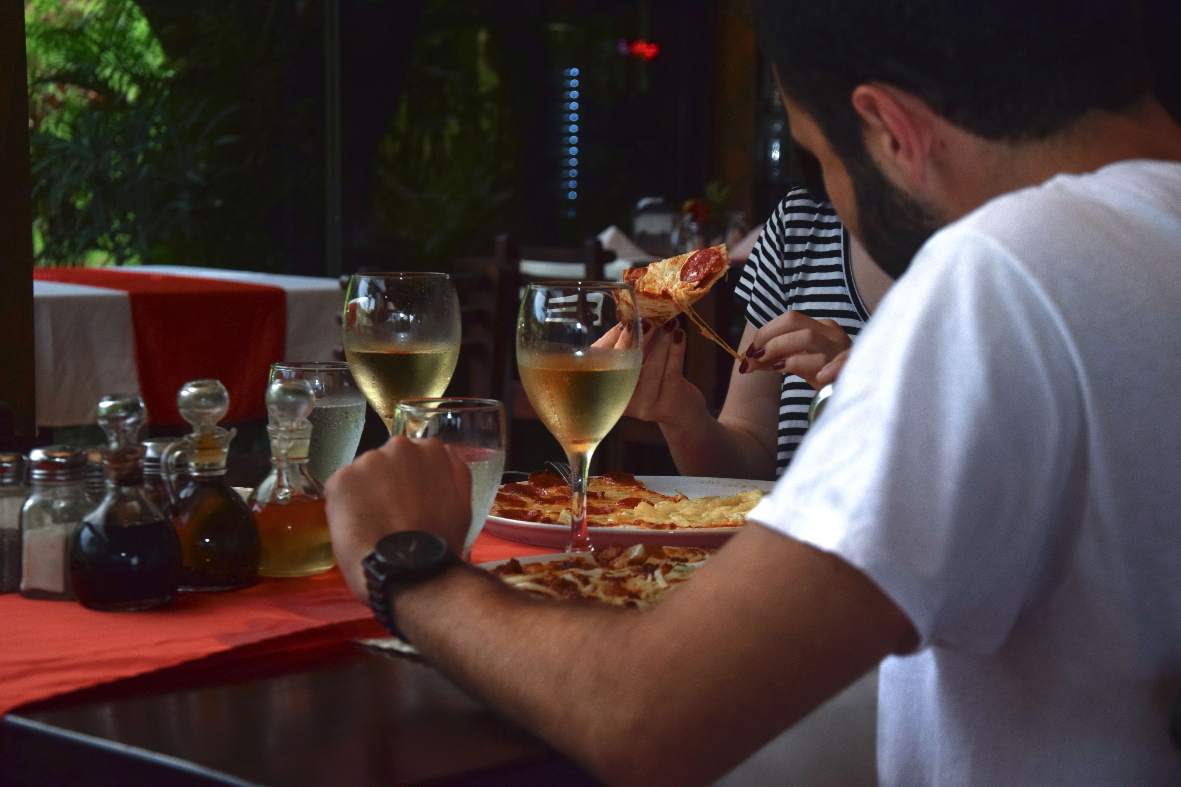 A person holding a slice of pizza at a restaurant table with wine glasses and condiments.