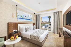 A hotel room with a large bed, a fruit bowl, a painting of sailboats, a TV, and a balcony with a view of palm trees.