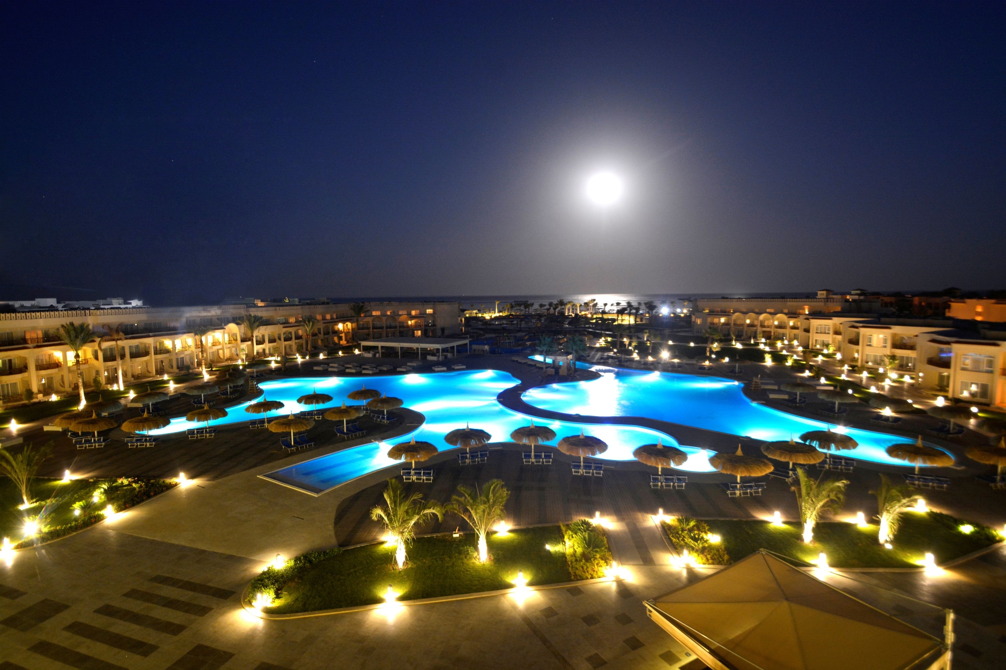 Une vue nocturne d’un complexe hôtelier avec une grande piscine, des palmiers et un croissant de lune dans le ciel.