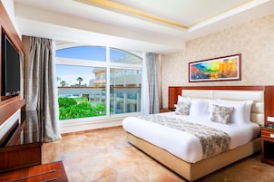 A hotel room with a large bed, a TV, a painting, and a view of greenery and buildings through the window.