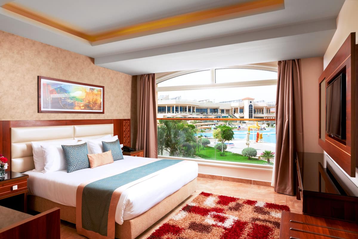 A hotel room with a large bed, a TV, a painting on the wall, and a view of a pool and buildings.