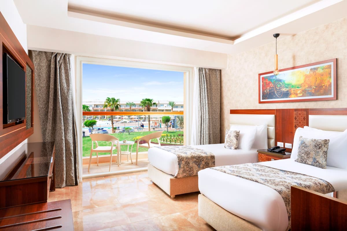 A hotel room with two beds, a TV, a balcony with a view of a resort and palm trees, and a painting on the wall.