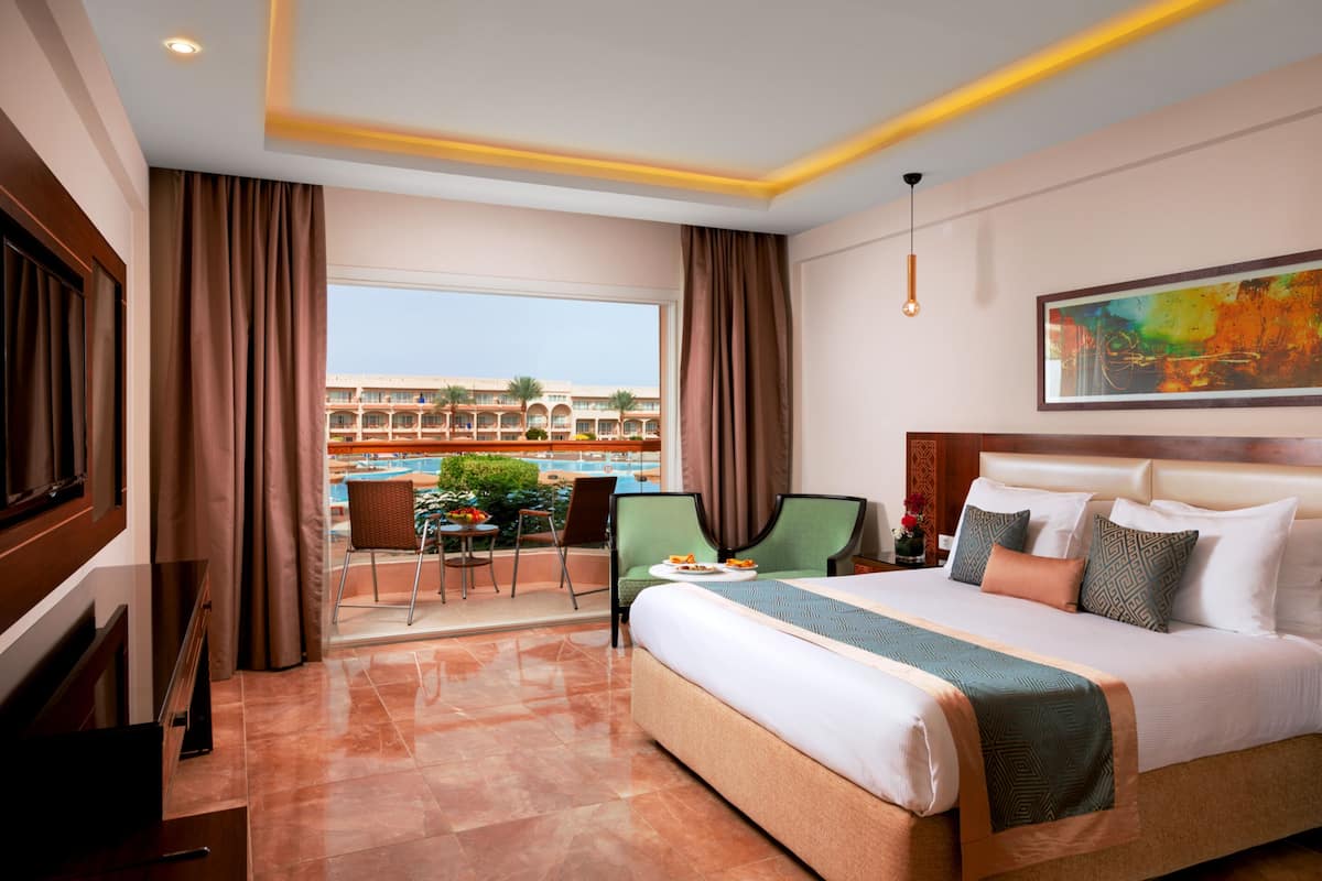 A hotel room with a large bed, a dining area with a table and chairs, and a view of a pool and building in the background.