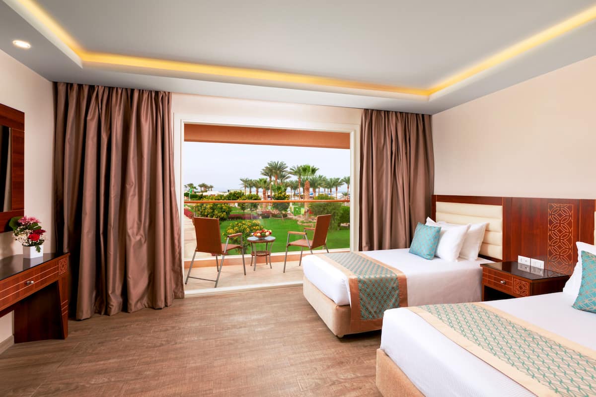 A hotel room with two beds, a desk, and a balcony with a view of a garden and palm trees.