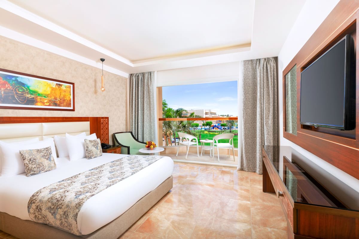 A hotel room with a large bed, a flat-screen TV, a balcony with a view of the city, and a dining area with chairs and a table.