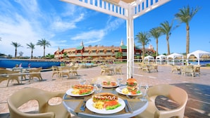 A dining area with outdoor tables and chairs, a pool, and a building with a red roof and multiple balconies in the background. - Pickalbatros Aqua Blu Sharm El Sheikh (Sharm El Sheikh)