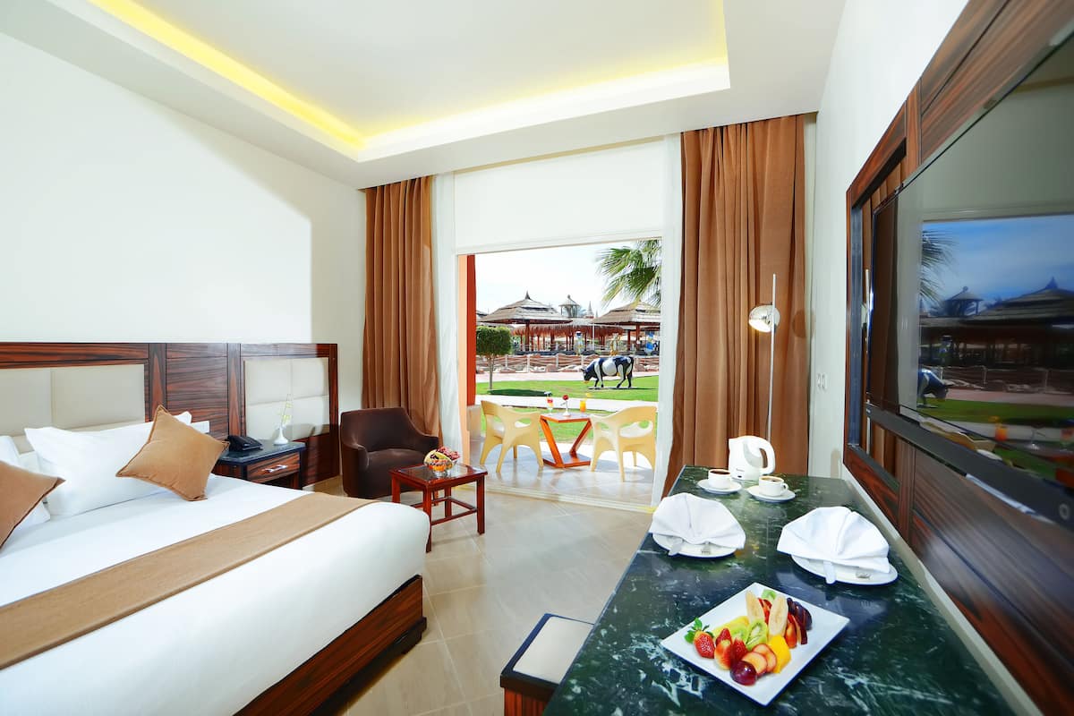 A hotel room with a bed, a desk with a fruit plate, a TV, and a view of a pool and outdoor seating area.
