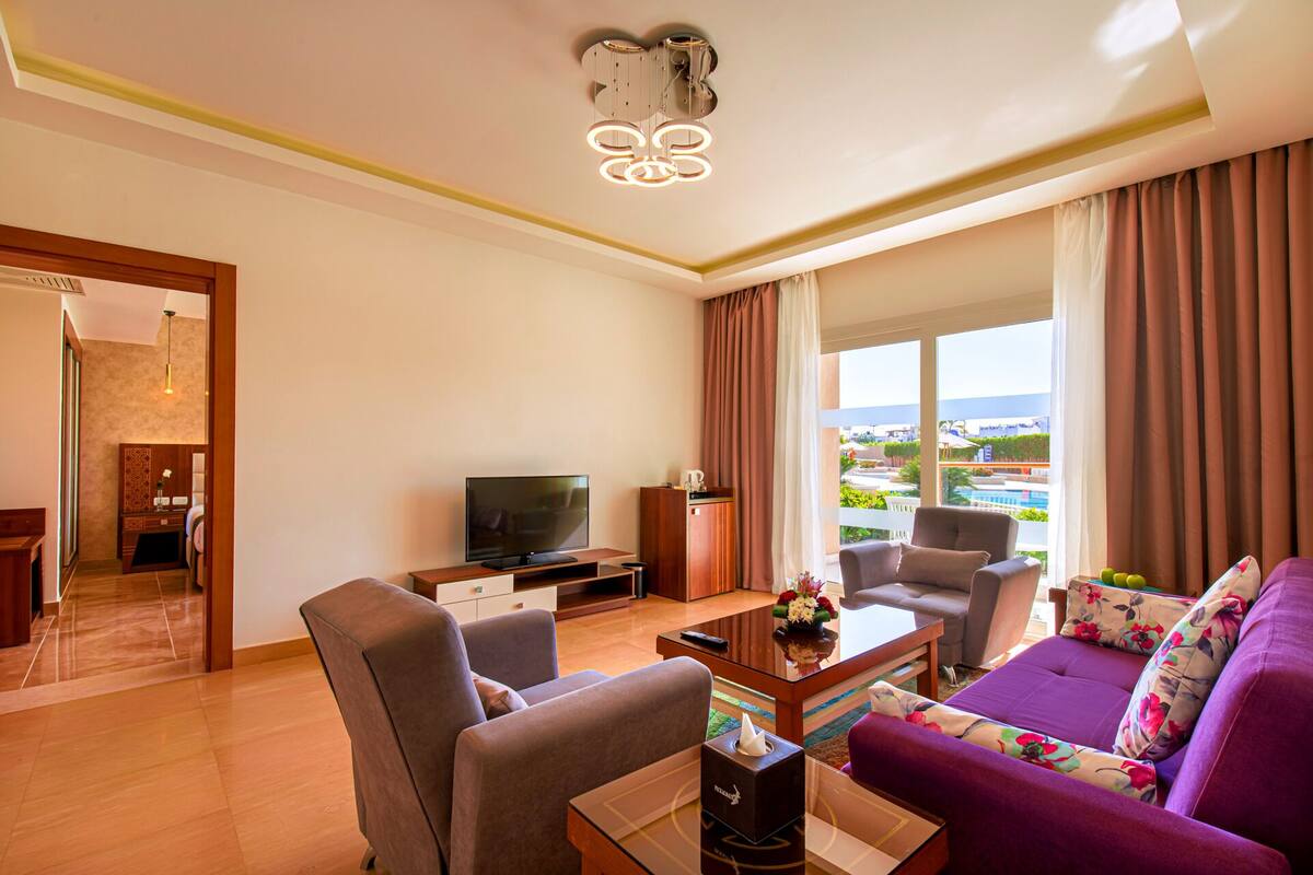 A modern hotel room with a sofa, armchairs, a TV, and a view of the city.