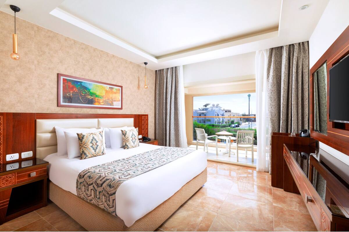 A hotel room with a large bed, a painting on the wall, a balcony with a view, and a TV.