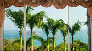 A view from a window with curtains, featuring palm trees and a distant island in the ocean.