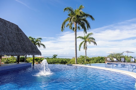 A swimming pool with a water fountain, surrounded by palm trees and a thatched-roof structure.