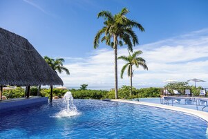 Una piscina con fuente de agua, rodeada de palmeras y una estructura con techo de paja.