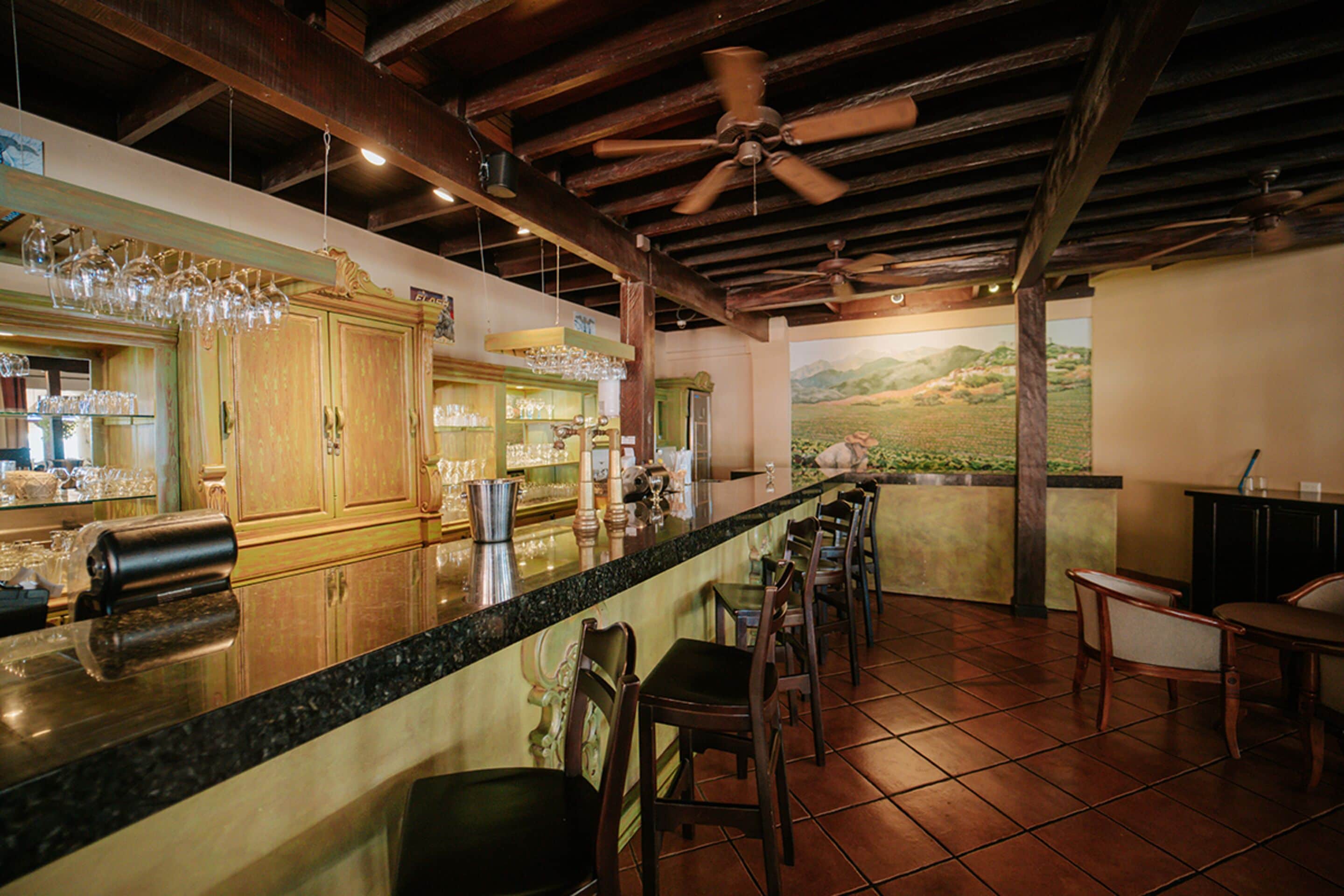 A bar area with a long counter, wooden chairs, and a ceiling fan.