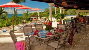 An outdoor dining area with tables set for two, surrounded by chairs, under a covered patio with red umbrellas.