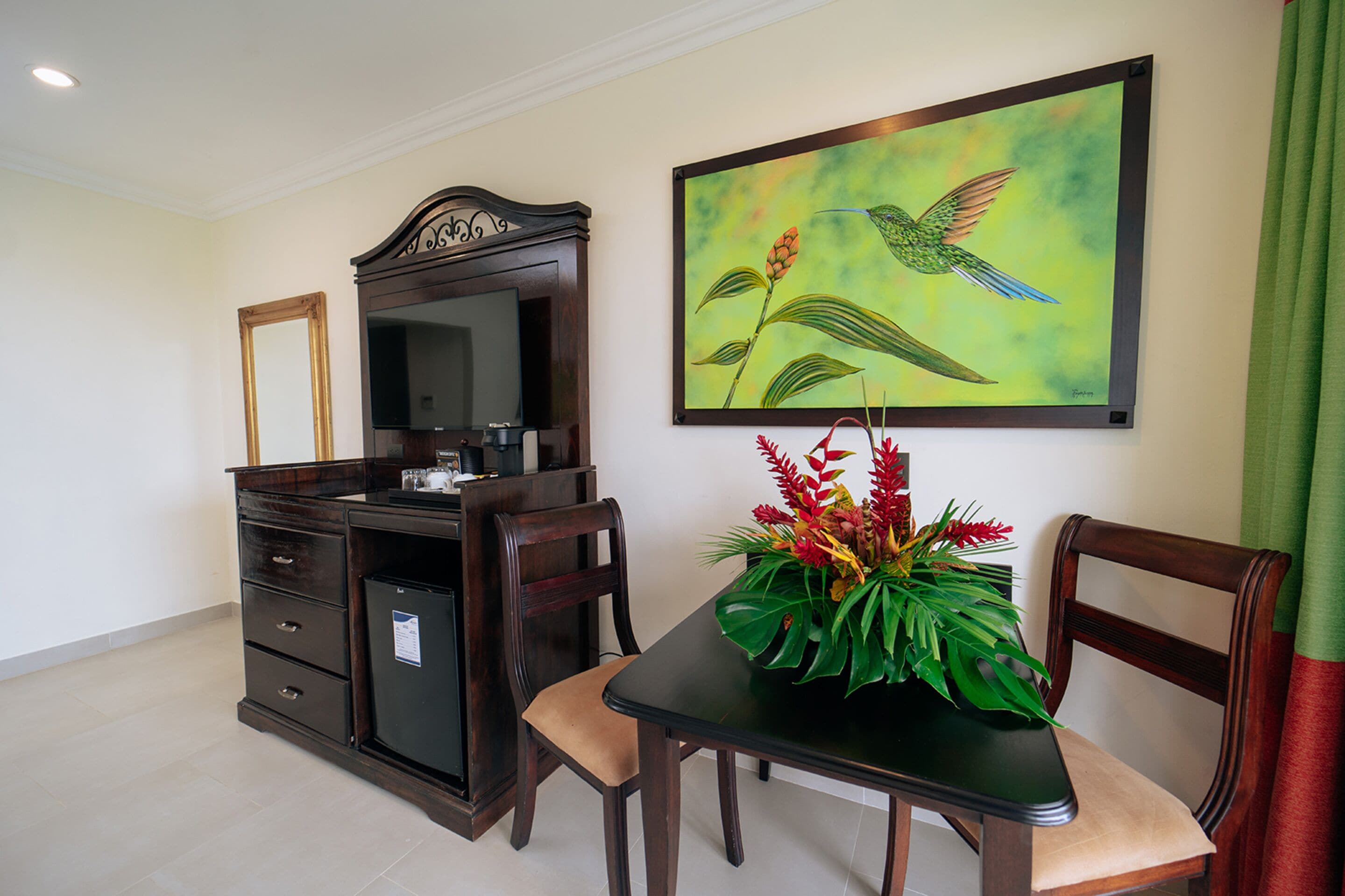Hotelkamer met een houten bureau, een stoel, een kleine koelkast, een spiegel en een schilderij van een kolibrie.