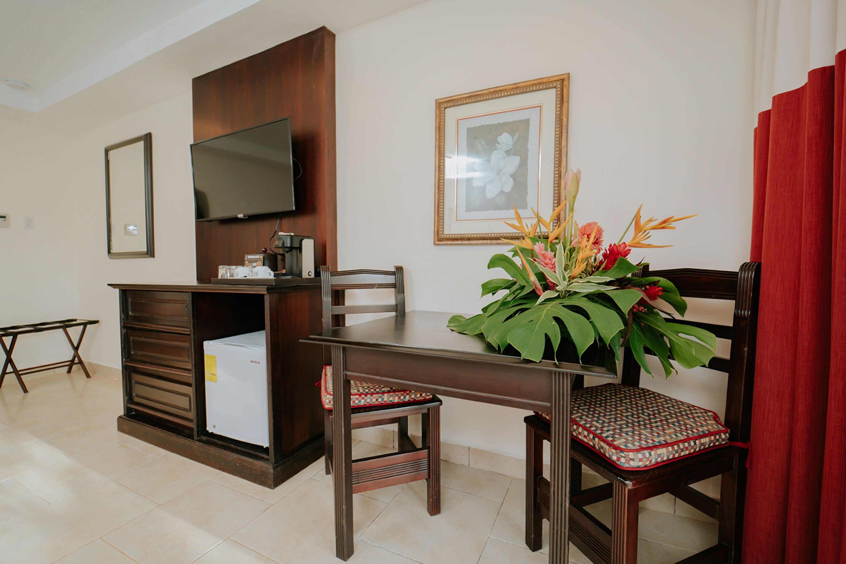 A hotel room with a wooden dining table, two chairs, a television mounted on the wall, a framed picture, and a potted plant.