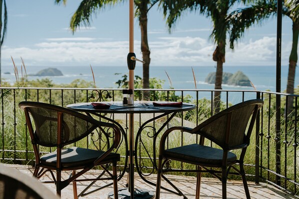 A patio with two wicker chairs and a table, overlooking a coastal view with palm trees and the ocean.