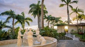 A fountain with statues, surrounded by palm trees and a stone pathway leading to a building with a tiled roof.
